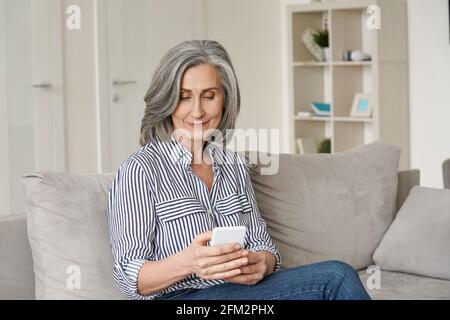 Happy mid age senior woman using smartphone sitting on sofa at home. Stock Photo
