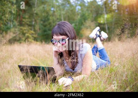 happy young woman with laptop on a sunny lawn. freelancer works in nature. student studies remotely on nature landscape outdoor Stock Photo