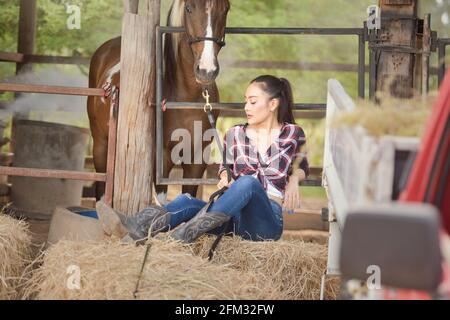 Beautiful woman sitting on a hay bale in a barn with her horse, Thailand Stock Photo