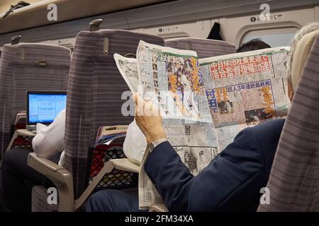 2 Japanese male commuters on a Shinkansen bullet train one reading work on his laptop and the other reading a newspaper baseball sports page, Japan Stock Photo