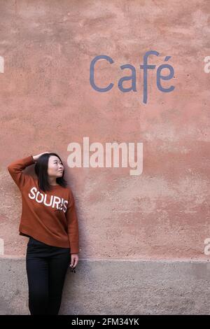 Asian woman leaning against a deep pastel pink sorbet coloured wall with a painted blue font café sign above her on the wall Stock Photo