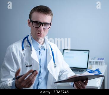 Young male doctor consults patient in hospital office, holding medical examinations in hands. General practitioner in white medical coat looks into Stock Photo