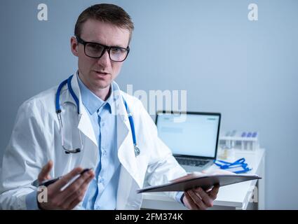 Young male doctor consults patient in hospital office, holding medical examinations in hands. General practitioner in white medical coat looks into Stock Photo