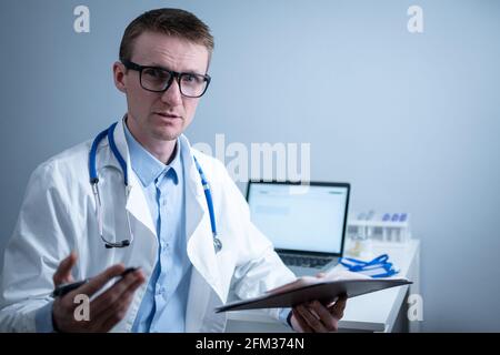 Young male doctor consults patient in hospital office, holding medical examinations in hands. General practitioner in white medical coat looks into Stock Photo