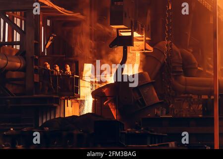 Molten iron pouring from ladle container into mold, workers control process, steel foundry factory, heavy metallurgy industry. Stock Photo