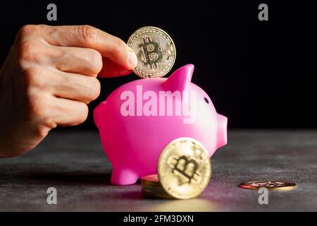 Concept image for using digital crypto currency for savings. A caucasian woman is putting a symbolic bitcoin (BTC) coin into a piggy bank as a saving Stock Photo