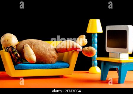 A quirky metaphorical concept image showing a potato man lying on a couch watching tv in a living room setting. Image for being couch potato, obesity, Stock Photo