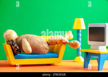 A quirky metaphorical concept image showing a potato man lying on a couch watching tv in a living room setting. Image for being couch potato, obesity, Stock Photo