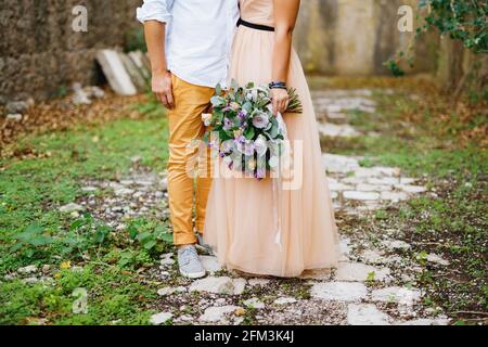 Half-portrait of groom embracing bride in a beautiful dress with a bouquet in her hands on the background of a stone wall Stock Photo