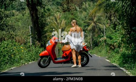 Woman on red scooter in white clothes drive on forest road trail. Dancing trip. One girl caucasian tourist in sunglasses dance, relax near motorbike. Stock Photo