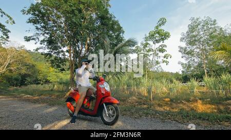 Man on red motorbike in white clothes drive on forest road trail trip. One men caucasian tourist go on scooter, nearby tropical palm tree. Stock Photo