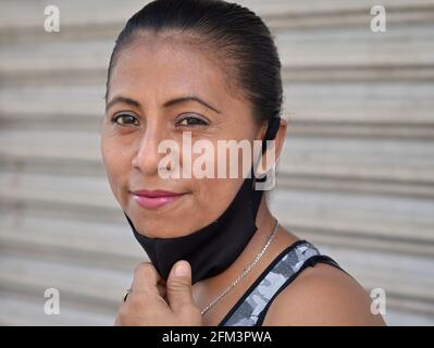 Middle-aged Mexican Latina woman with beautiful brown eyes pulls a non-medical black fabric face mask down during the global coronavirus pandemic. Stock Photo