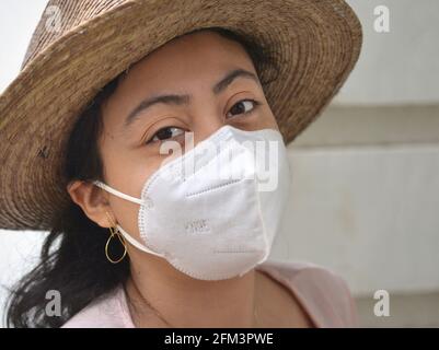 Brown-eyed young Mexican woman wears a worn Panama summer straw hat and a white KN 95 protective face mask during the global coronavirus pandemic. Stock Photo