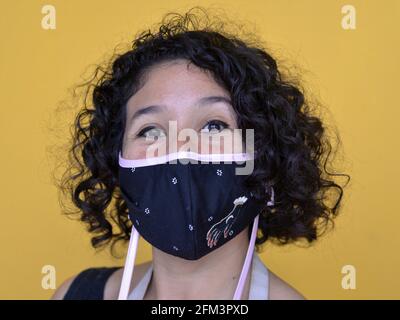 Young Mexican woman with corkscrew curls wears stylish non-medical cloth face mask during the global coronavirus pandemic and looks at camera. Stock Photo