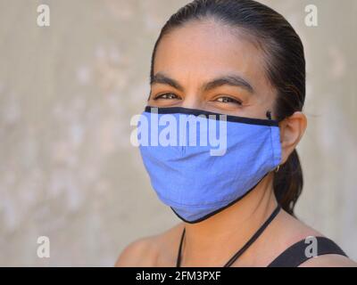 Resourceful Mexican latina woman with smiling eyes wears a blue non-medical face mask during the global coronavirus pandemic and poses for the camera. Stock Photo