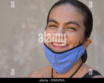 Cheerful Mexican woman with slicked back long hair pulls her blue non-medical face mask down and smiles for the camera. Stock Photo
