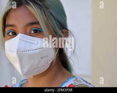 Mexican teen girl with dyed hair, big brown eyes and elaborate blue eye makeup wears a white KN95 face mask during the global coronavirus pandemic. Stock Photo