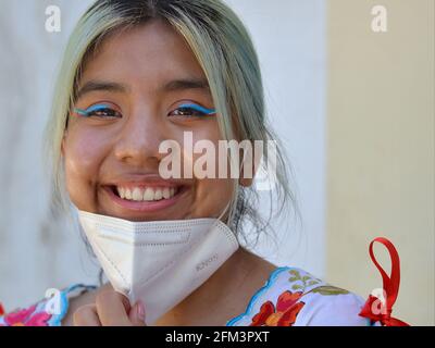 Cheerful Mexican teen girl with elaborate blue eye makeup pulls down her white KN95 face mask and smiles for the camera during coronavirus pandemic. Stock Photo
