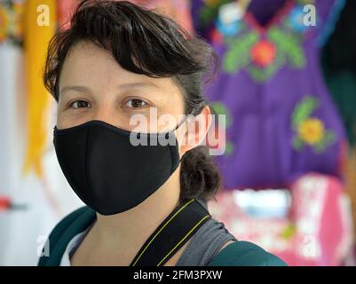 Young Mexican woman wears a black face mask during the global coronavirus pandemic and poses for the camera in front of a folkloric clothes stall. Stock Photo