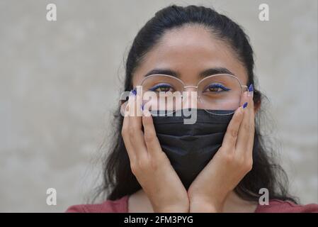 Young stylish beautiful Mexican woman with blue eyeliner and blue nail enamel wears eyeglasses and a black pandemic face mask and looks at the viewer. Stock Photo