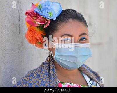 Young chubby Mexican Yucatecan woman with colorful flowers in her hair wears a light-blue surgical face mask during the global coronavirus pandemic. Stock Photo