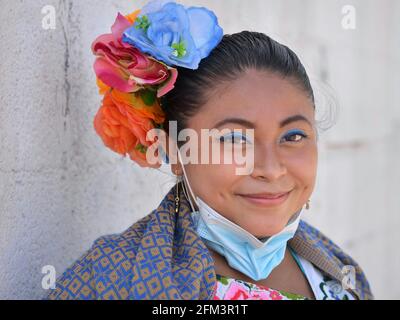 Chubby young pretty Mexican Yucatecan woman with colorful flowers in her hair pulls down her light-blue surgical face mask and smiles with her eyes. Stock Photo