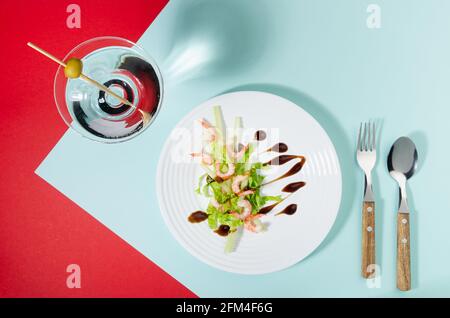 Colorful organic salad of shrimps, greens, celery, balsamic sauce with cocktail decorated green olive on red, minty background, flat lay. Modern conce Stock Photo