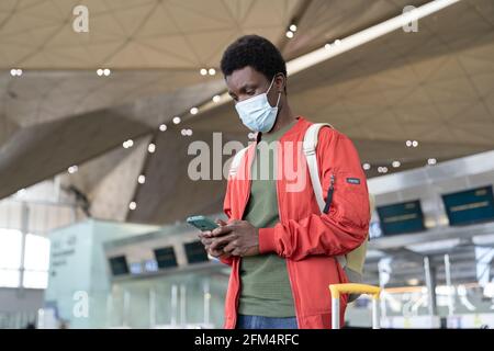 Young black male traveler in airport wait for flight use smartphone wear mask in covid-19 epidemic Stock Photo