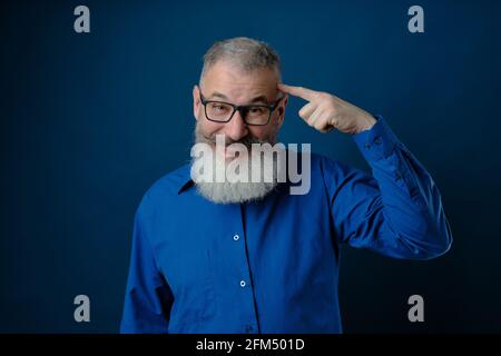 Bearded middle age man dressed blue shirt pointing to head with forefinger, great idea or thought concept, good memory, blue backdrop Stock Photo