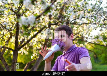 Defocus portrait of handsome caucasian man with cone flowers in lilac garden Funny cheerful young brunette guy smelling fresh flowers and looking at Stock Photo