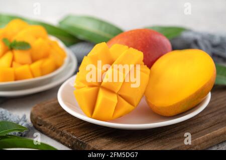 Diced fresh mango fruit on a white plate over wooden cutting board with leaves on gray table background. Stock Photo