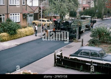 A tarmac (asphalt) machine and workers resurfacing a suburban street in London, England, UK in 1984. A truck, from a company named Bardon, has delivered a batch of fresh material to the tarmac laying vehicle. The new surface is evident behind the machinery. The street is a typical of London’s outer ‘Metroland’ suburbs with houses mostly semi-detached and dating from the 1930s/40s. This image is from an old amateur 35mm Kodak colour transparency – a vintage 1980s photograph. Stock Photo