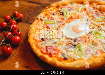 Close up view of spicy italian pizza with egg, ham, cheese and tabasco pepperoni on wooden table with decorating cherry tomatoes. Stock Photo