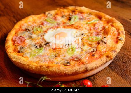 Close up view of spicy italian pizza with egg, ham, cheese and tabasco pepperoni on wooden table stock photo. Stock Photo