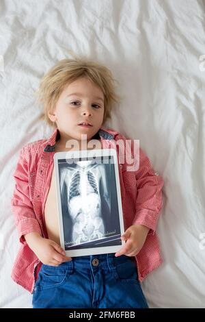 Toddler child, blond boy, holding x-ray picture on tablet of child body with swallowed magnet showing, child swallow dangerous object Stock Photo