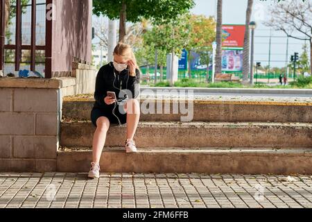 Woman in sportswear and face mask looks at her phone sitting on stairs Stock Photo