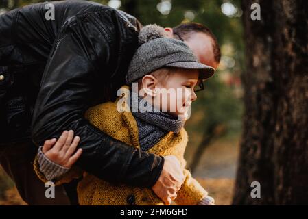 Father and son having hug in autumn city park. Happy kid walking among fallen leaves. Kids fashion. Boy wearing trendy yellow coat, cap and scarf. Smiling young boy outdoors. Kid jumping and Stock Photo