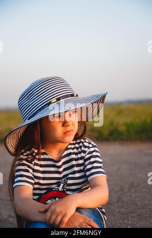 Close up portrait of little sad girl in blue and white striped sun hat and shirt sitting alone at the road Stock Photo