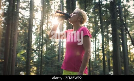Middle shot of a Caucasian woman in sportswear standing in the forest, drinking water from a bottle Stock Photo