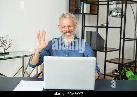 Cheerful contemporary senior grey hair businessman sits in front of laptop and waving hello into webcam, greeting colleagues or business partners involved video meeting, using app for video connect Stock Photo