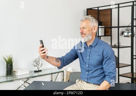 Mature grey haired businessman in smart casual wear is using smartphone for video connection sitting in relaxed pose at the office, holding meeting with business partners, talking with colleagues Stock Photo