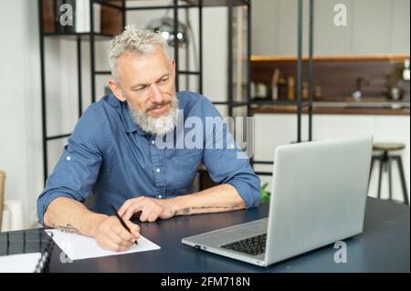 Modern middle aged bearded tattooed man working with a laptop in home office, senior grey hair male in smart casual shirt watching webinar and writes down, takes notes with a pen, planning schedule Stock Photo