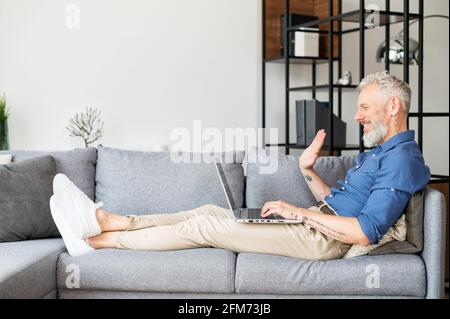 Side view mature bearded man talking online via video connection on the smartphone, modern grey-haired male lying down on the couch in relaxed pose and waving into webcam. Video call concept Stock Photo