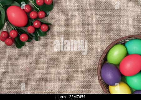 Multicolored Easter eggs in a wicker basket and red berries on a burlap background. Stock Photo