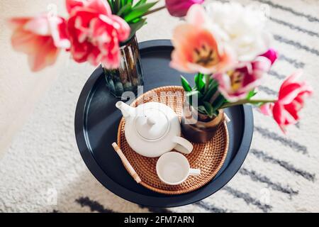 Interior and home decor. Fresh tulips flowers put in vases on coffee table with tea kettle and cup on rattan tray. Top view Stock Photo