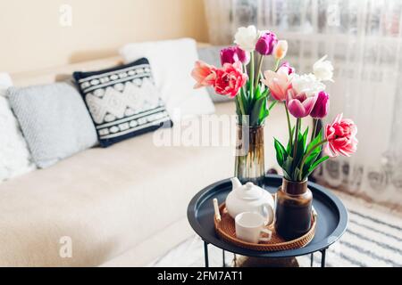 Interior and home decor. Fresh tulips flowers put in vases on coffee table with tea kettle and cup on rattan tray. Cozy living room Stock Photo