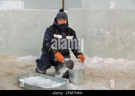 A worker assembler in winter overalls puts wet snow from a trough into a metal bucket Stock Photo