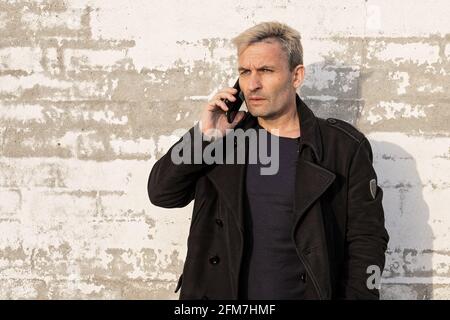 Attractive blonde middle aged man is posing with mobile phone against white brick wall. Male modelling, fashion photoshoot, professional. Front view. Stock Photo