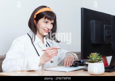 A female doctor wearing a headset makes an online video call with a patient and writing something on clipboard, prescription, paperwork, patient check Stock Photo