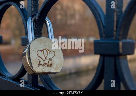 Semi-circular shiny golden-silver padlock with small spots left by time and engraved with two woven hearts fixed on round elements of bridge fence. Co Stock Photo